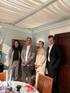 A group poses beside a dining setup in a warmly lit room with soft blue walls, Greek key molding and a fabric-draped ceiling, creating an intimate and elegant ambiance.