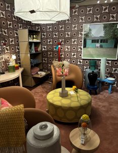 Retro meets playful in a room of earthy tones and rounded forms. Graphic wallpaper, sculptural ceramics, and soft furnishings create a layered, inviting scene anchored by bold seating and an oversized drum pendant.