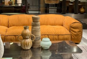 Bax features a plush silhouette in warm ochre velvet, with deep channels and rounded arms. Paired with handcrafted ceramic vases and a sculptural table, it exudes retro charm and casual luxury in a contemporary lounge setting.
