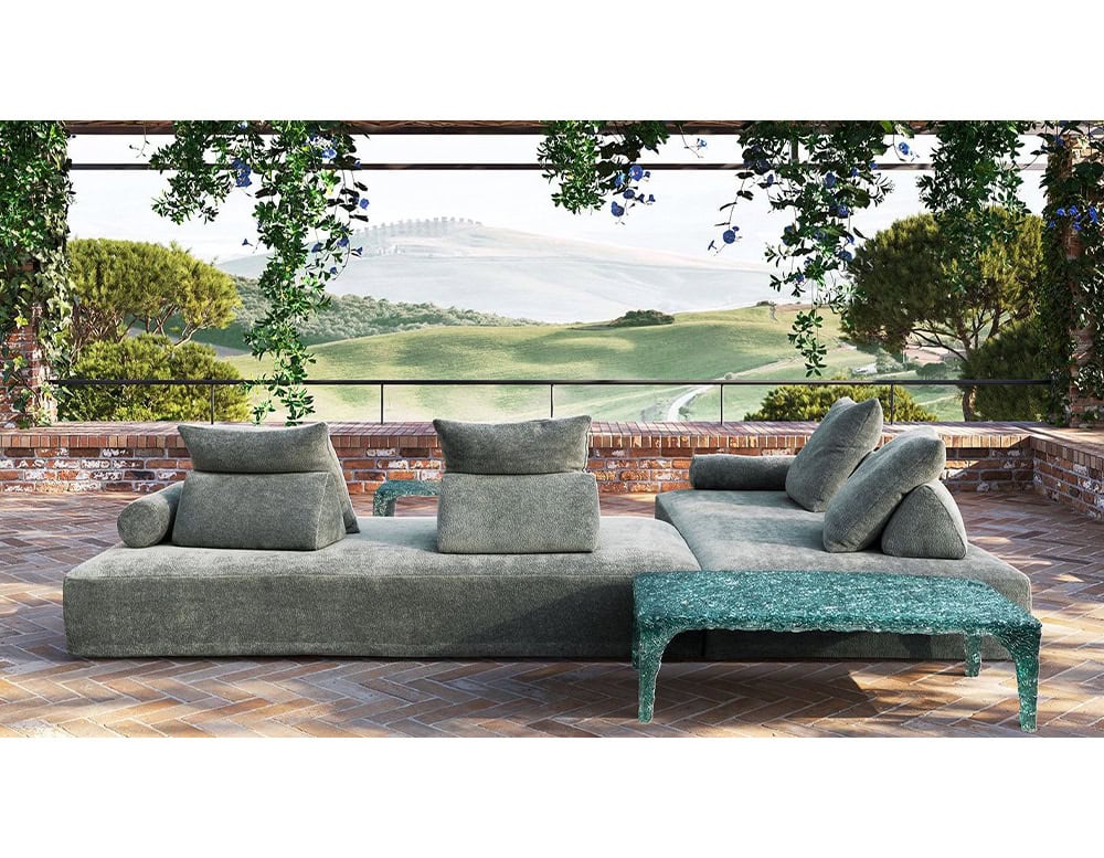 Low-slung modular seating in textured green upholstery with angled backs and cylindrical bolsters. Shown on a terrace with terracotta brick flooring and scenic countryside views framed by hanging foliage.