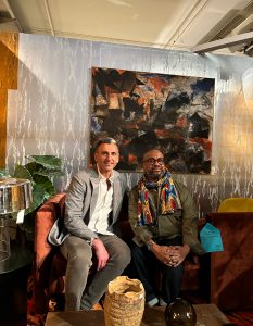 Two guests sit on a deep orange sofa beneath a striking abstract painting, surrounded by bold textures and curated modern decor.