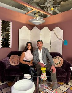 Two guests are seated on a velvet sofa in a chic, artfully staged showroom vignette with sculptural decor, playful pillows, and fresh flowers on the coffee table.