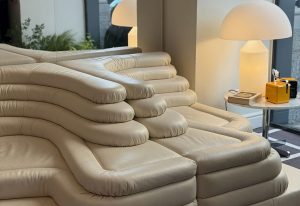 Sculptural sectional in cream leather with layered, contoured cushions. Its flowing form and plush texture create a striking lounge piece, paired with soft lighting and a modern side table.