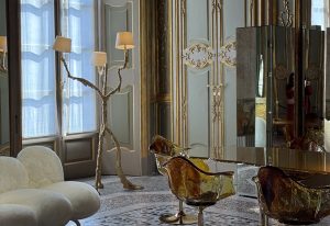 Amber-toned chairs shimmer around a mirrored table. Gilded wall panels and a sculptural floor lamp create regal contrast. Velvet, glass, and ornate trim blend into a luxe interior. Refined materials and bold forms define this harmonious setting.