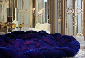 In a gilded interior, the indigo velvet lounge sprawls in voluptuous, braided forms. Its plush texture contrasts the ornate backdrop of gold-trimmed panels and mirrored walls. Regal and modern, it dominates the space with tactile drama.