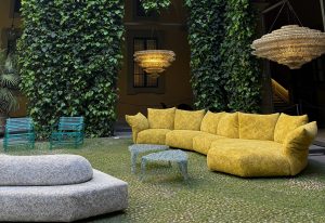 Vibrant outdoor setting with sculptural seating and soft geometric lines. A golden curved sofa anchors the space, paired with terrazzo-style tables and glowing chandeliers suspended above a lush green courtyard.