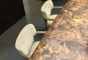 Henning showcases a rich, organic wood grain tabletop with intricate natural patterns. Paired with soft upholstered armchairs, it merges comfort and craftsmanship in a refined, modern setting.