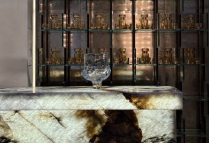 Henning features a bold stone bar front with dramatic veining, paired with a sculptural glass vessel. Behind, shelves of glass decanters create a luminous, golden backdrop that adds depth and elegance.
