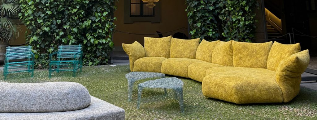 Vibrant courtyard scene. A mustard-textured modular sofa anchors the view, framed by living green walls and glowing chandeliers. Sculptural tables and emerald seating add charm to this lush, elegant installation.
