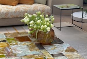 Multicolored mosaic glass table, topped with a warm amber vase of fresh white tulips. A floral-upholstered sofa with ochre pillows and minimal black-framed side tables complete the cozy, elegant setting.