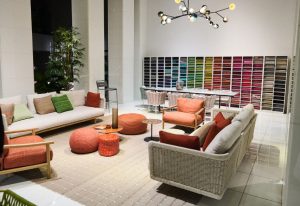 Colorful fabric swatches line the back wall of this bright, modern showroom. Light-toned woven sofas and lounge chairs with vibrant cushions are gathered around playful poufs and tables, grounded by greenery and a sculptural, branching chandelier.