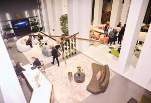 Overhead view of a vibrant showroom event filled with sculptural lighting, modular seating areas, and polished surfaces. Neutral tones meet bright accent furniture, while guests mingle and explore the dynamic, multi-zone environment.