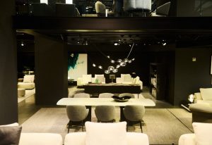 A dramatic two-level showroom with moody lighting and modern appeal. Plush cream seating contrasts with dark architectural details, while a floating chandelier and artful accents add sculptural interest above sleek dining and lounge arrangements.