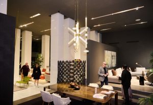 A dynamic showroom with soaring ceilings, glossy columns, and sculptural lighting. A long wood table anchors the scene, with elegant decor, cozy seating areas, and guests engaged in relaxed conversation throughout the sophisticated space.