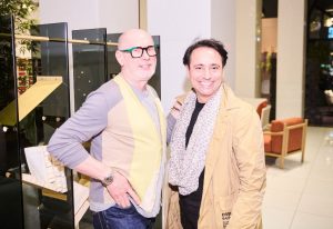 Two guests stand and smile inside a bright showroom space, surrounded by modern glass shelves and warm-toned armchairs.