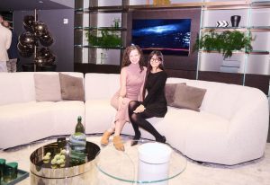 Two guests are seated on a curved cream sofa in a modern showroom, framed by ambient lighting, greenery, and minimalist shelving.