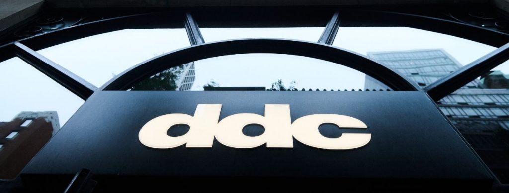 A close-up view of the DDC storefront sign, photographed from below with a city skyline reflected in the large arched window above.