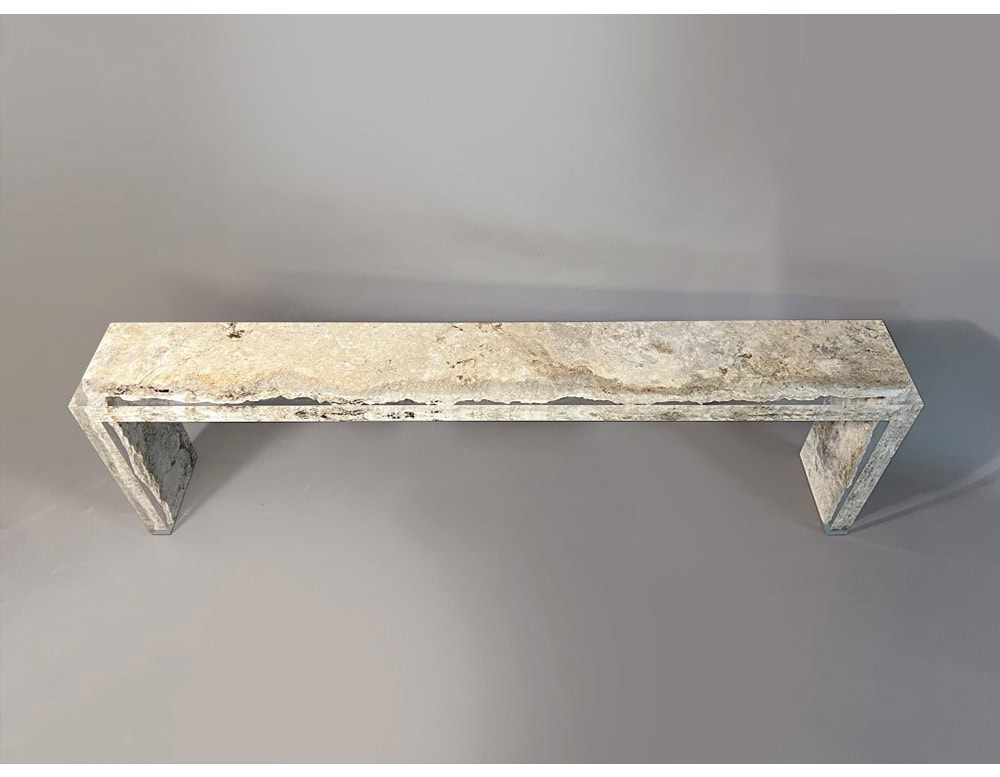 The Rapolano Console Table showcases a long travertine slab with raw, textured edges. Its minimalist structure highlights the natural beauty of the stone, supported by sleek geometric legs.
