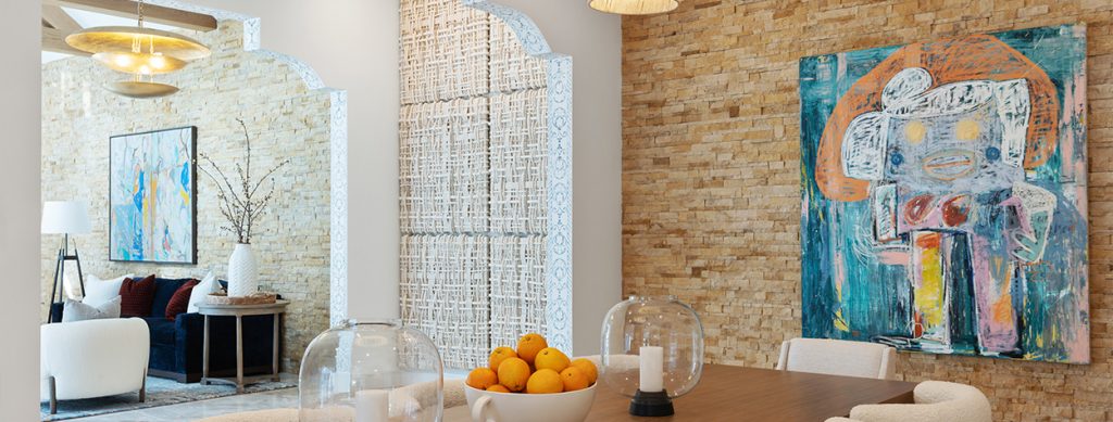 A contemporary dining area features a wooden table with a bowl of oranges and glass candle holders. Behind it, a textured wall displays a colorful abstract painting with playful, childlike strokes.