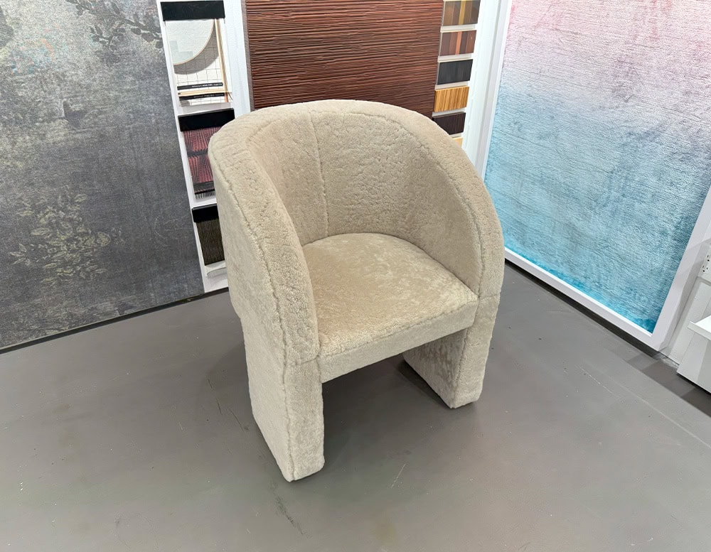 Bax Lazybones Armchair features a cozy fully upholstered form with curved arms and a plush seat in a pale cream tone. It rests on a muted gray floor, surrounded by decorative wall panels that hint at a materials display setting.