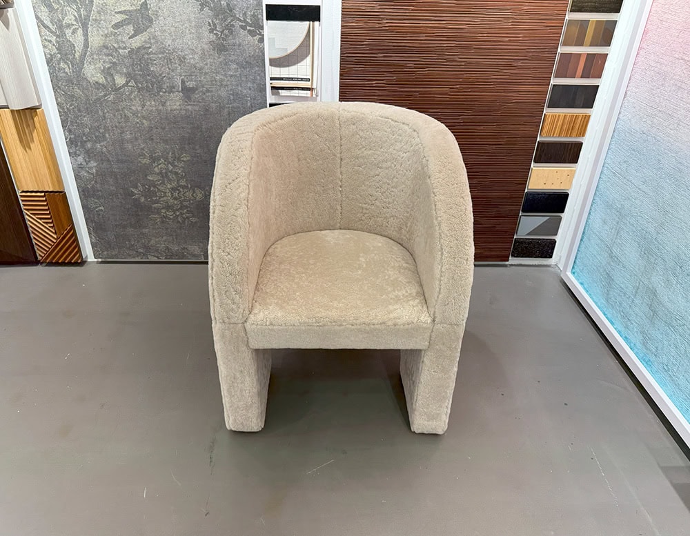 Bax Lazybones Armchair offers a soft plush presence with fully upholstered arched arms and a rounded backrest. Shown here in a creamy tone, it sits on a neutral floor, framed by contrasting textures and color swatches in the background.