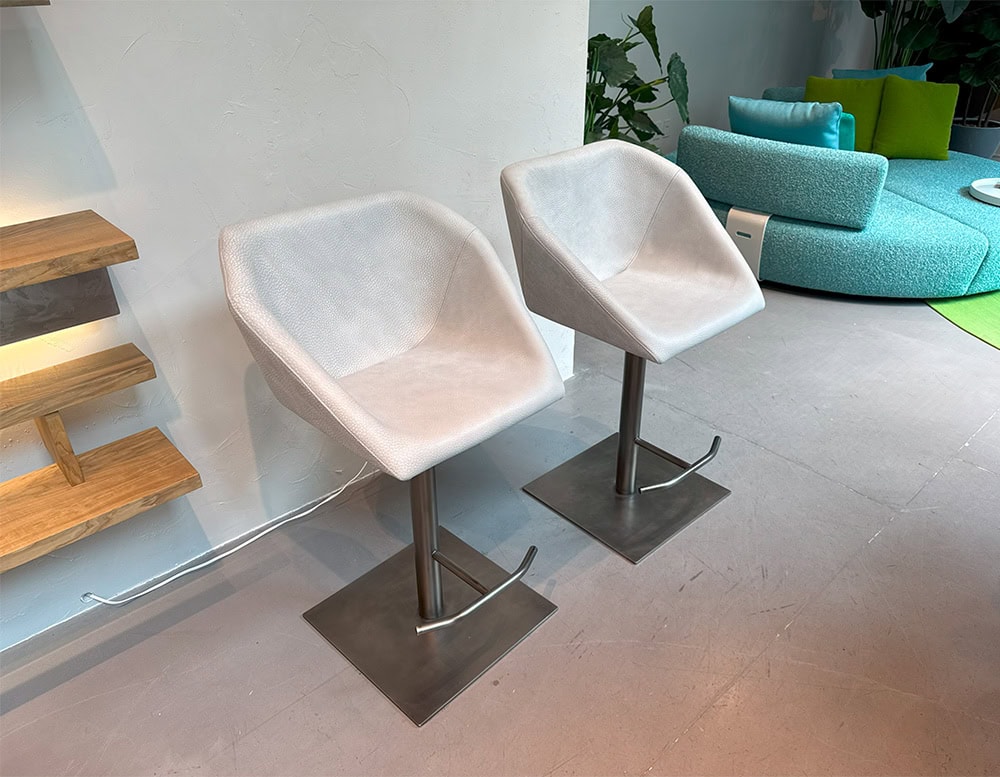 The Hen Hexagon Stools are shown in a pair, upholstered in white fabric with a subtle texture. Each rests on a brushed metal pedestal with footrest, set against wood shelving and a vibrant turquoise lounge in the background.