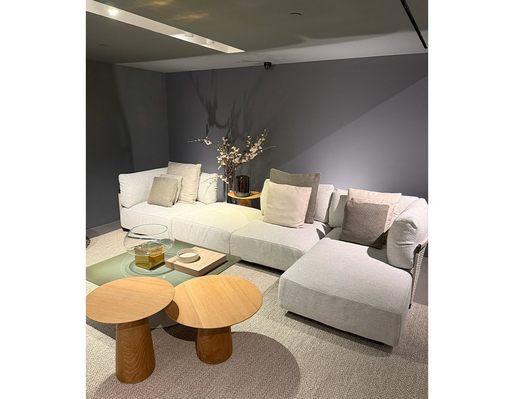 The Tri Nodi Grouping sectional anchors a cozy corner with soft cushions and neutral fabrics. Two rounded wood side tables and a square green coffee table create a layered, modern lounge setup accented with delicate floral decor.