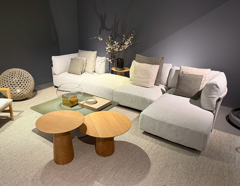 The Tri Nodi Grouping sectional is arranged with neutral-toned cushions and accompanied by a pair of rounded wood side tables. A green square coffee table, decorative tray, and glass vase complete the relaxed, modern lounge setting.