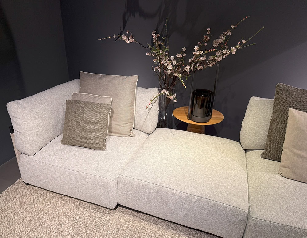 The Tri Nodi Grouping sectional is styled with soft neutral cushions and paired with a round wooden side table. A black lantern and delicate floral arrangement complete the serene lounge setting against a dark wall.