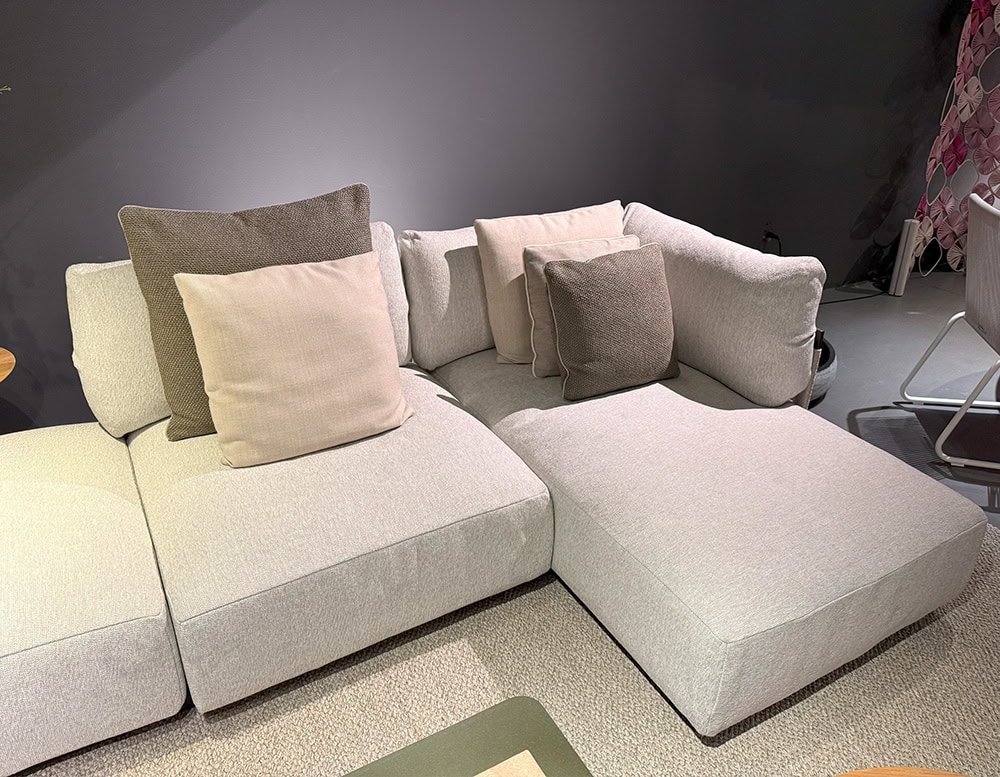 The Tri Nodi Grouping sectional sofa is arranged in an L-shape with soft beige upholstery and contrasting taupe and ivory pillows. The design offers a relaxed, modular lounge setup on a textured neutral rug.
