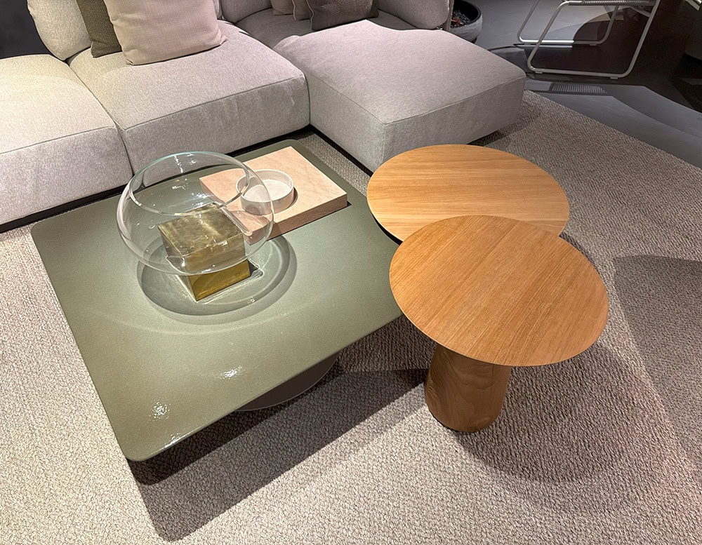 The Tri Nodi Grouping presents a mix of forms and textures. A square olive green coffee table with a glass and brass sculpture sits beside two overlapping round wood tables, all placed on a soft beige rug next to a modular sofa.