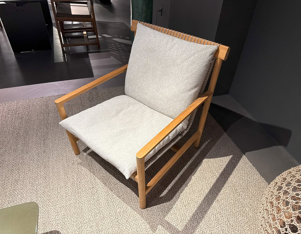 The Tri Nodi Armchair features a solid wood frame with simple, linear construction. Its light gray cushions are plush and relaxed, adding softness to the structured design. The chair rests on a textured beige rug within a warm, modern lounge.