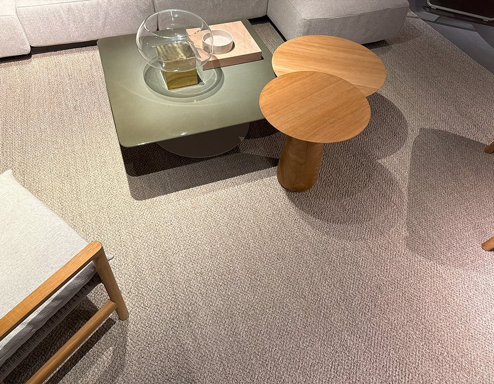 The Tri Nodi Grouping features two overlapping wooden tables with rounded tops and sculptural bases, paired with a low matte green coffee table. The arrangement rests on a textured beige rug, creating a warm and balanced lounge setting.