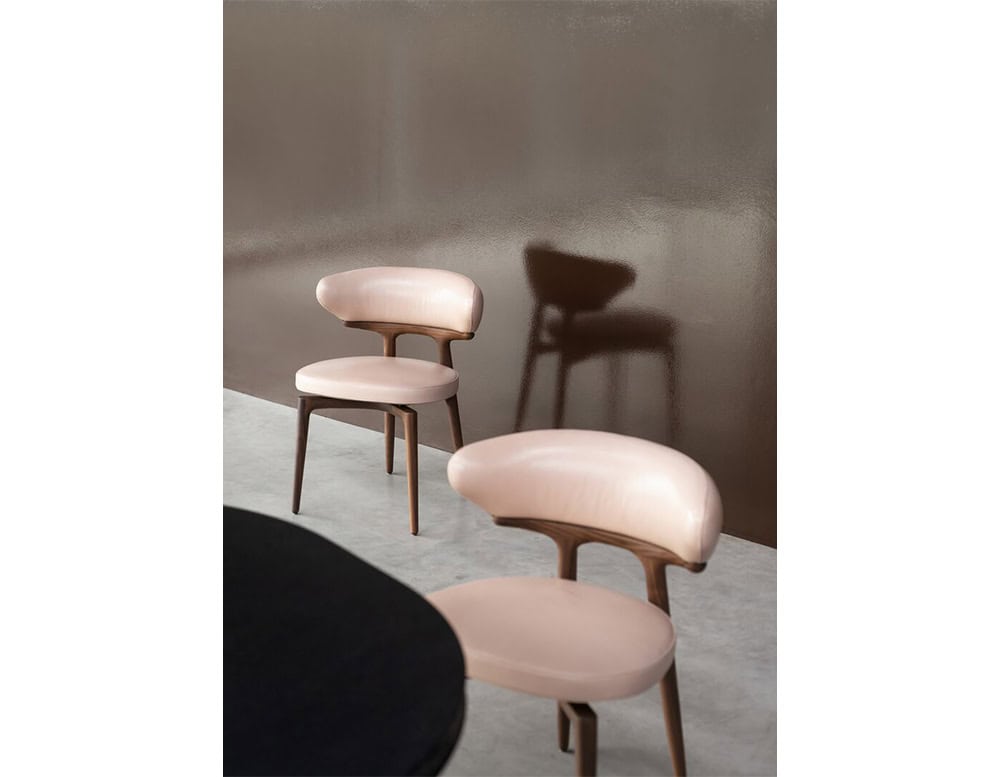 The Bax Clotilde Chair exudes subtle elegance with its blush-toned upholstery and sculpted wood base. Set against a sleek wall, the chairs create a refined yet inviting dining atmosphere.