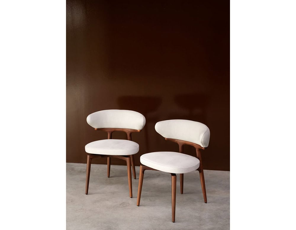 The Bax Clotilde Chair features soft ivory upholstery and a sculptural walnut base. Styled in pairs against a warm backdrop, it creates an atmosphere of understated luxury.