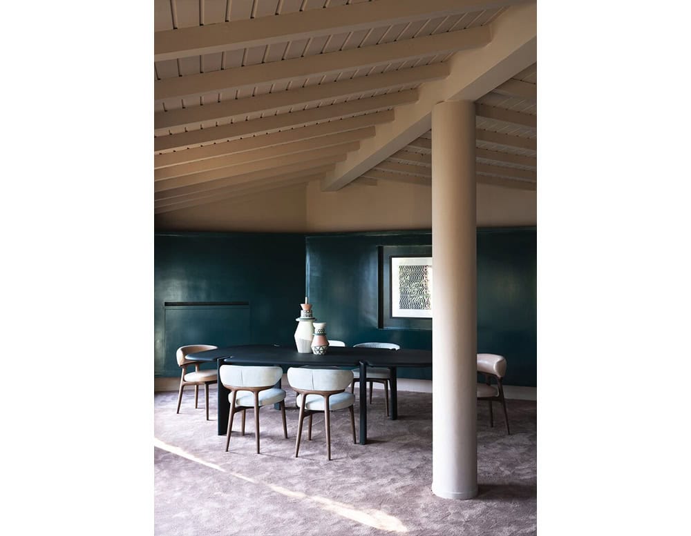 The Bax Clotilde Chair surrounds the table with elegant curves and soft neutral tones. Its sculpted wood frame complements the architectural ceiling and bold teal walls for a harmonious dining space.