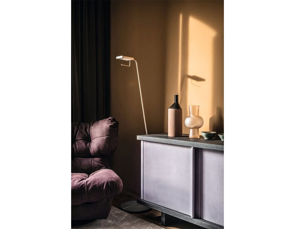 A soft-paneled sideboard from the Bax Joni Collection is shown in a muted lavender tone, paired with a deep brown textured top. Beside it, a plush armchair and a minimal floor lamp complete the corner arrangement.