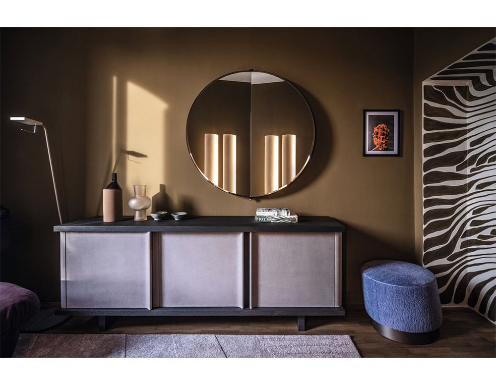 A Bax Joni sideboard with softly paneled fronts and a dark textured top is placed against an ochre wall. A round mirror reflects vertical lights, while a sculptural pouf, a graphic art piece, and a zebra-patterned niche add layered visual interest.