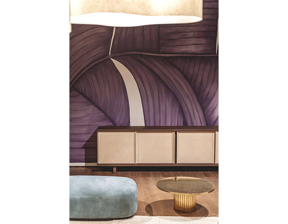 A Bax Joni sideboard with soft neutral panels and a dark top is positioned against a bold mural wall in deep violet tones. In the foreground, a low, light blue pouf and a sculptural round table with a fluted brass base complete the setting.