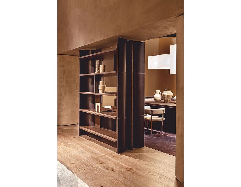 This view features a Bax Joni bookcase in a dark wood finish, used as a room divider. Its sculptural side panels and clean shelves create a functional yet architectural statement, while the neutral décor and soft lighting complete the warm setting.