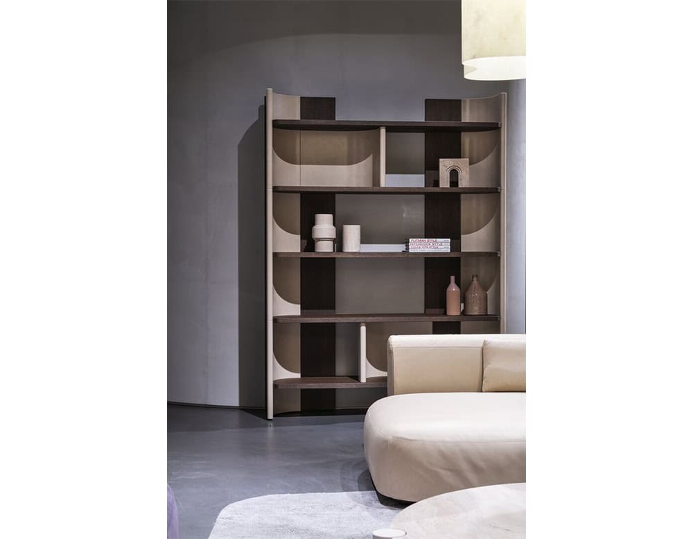 The image highlights a version of the Bax Joni bookcase in a lighter finish with contrasting dark accents. The open structure is styled with minimal ceramic and decorative objects, while a soft, curved seating piece appears in the foreground.