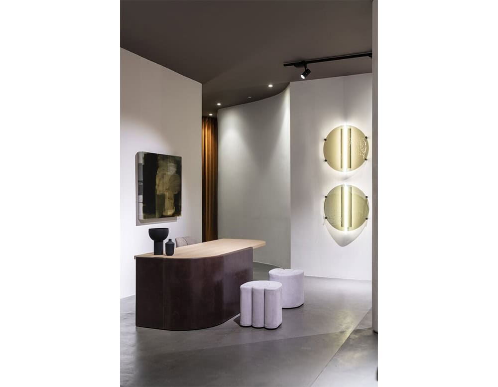 A Bax Joni console with a softly curved base and light wood top, paired with two cylindrical stools and accented by sculptural wall mirrors and a monochrome painting.