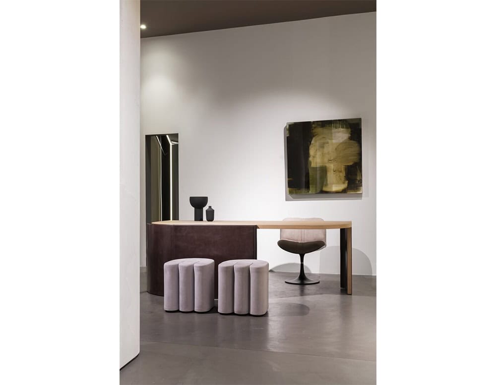 A Bax Joni console with a rounded base and light wood top, accompanied by two cylindrical stools and a swivel chair. A dark-toned artwork and minimalist vases complete the setting.