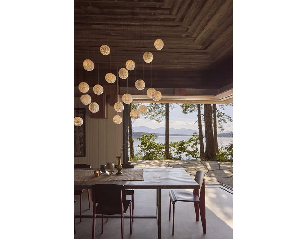 The Boc 118 Series chandelier creates a dramatic cloud of glowing glass spheres, suspended over a dining table with a serene lakeside view, blending sculptural lighting with natural surroundings.