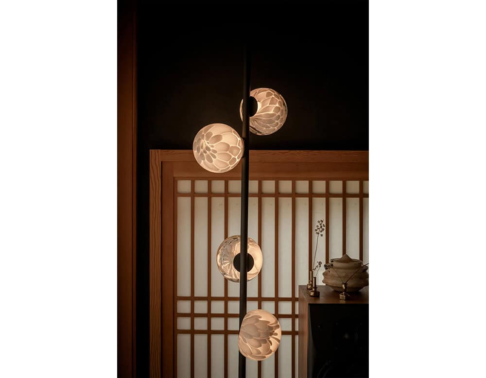 The Boc 118 Series floor lamp glows softly with hand-blown glass spheres along a dark vertical frame, creating a warm sculptural presence in a serene, wood-accented space.