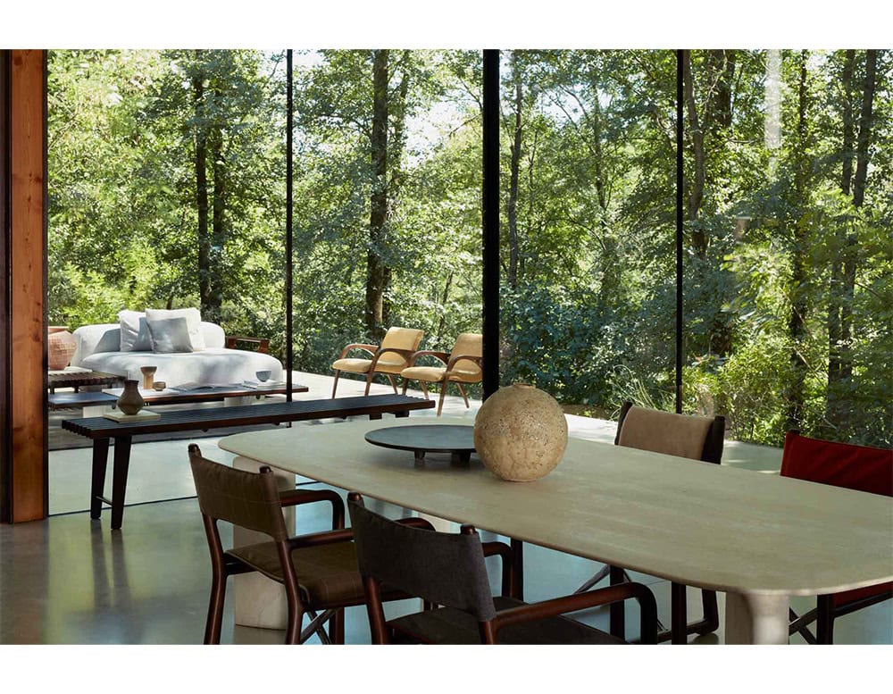 The Ext Elba Coffee Table Outdoor enhances the architectural setting with its refined slatted wood design, elegantly complementing both dining and lounge areas surrounded by serene nature.