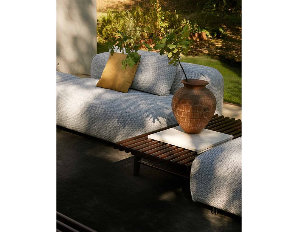 The Ext Elba Coffee Table Outdoor integrates seamlessly into open-air lounging spaces, featuring a warm wooden slat surface that pairs effortlessly with plush seating and natural textures, creating a relaxed yet sophisticated setting.