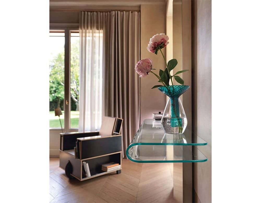 The Fiam Let Me See Mirror features a refined glass shelf with curved edges, creating a light and elegant focal point. A delicate vase with pink flowers adds a soft, organic accent to the scene.