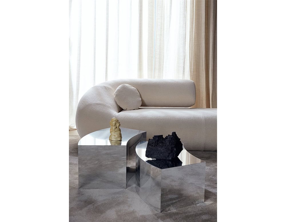 A pair of mirror-polished stainless steel coffee tables with curved forms reflect their surroundings, adding sculptural contrast to the soft, cream upholstered sofa.