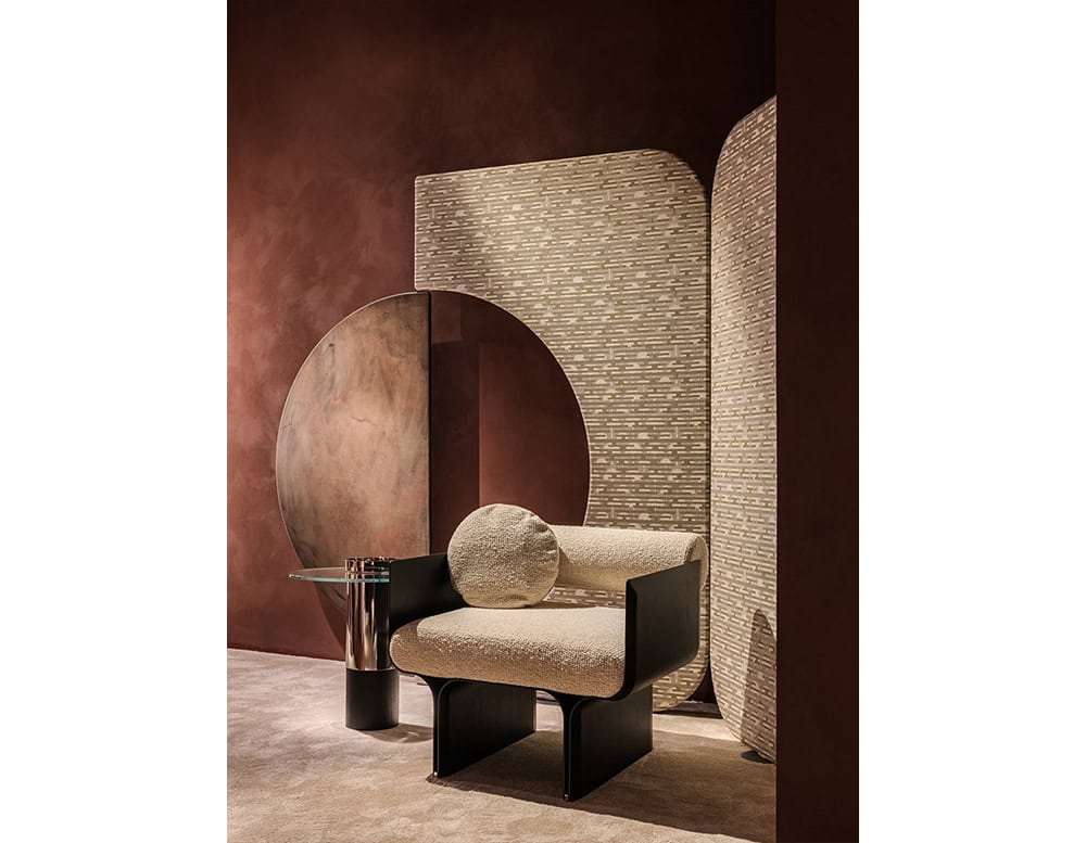 The Enso Screen presents a curved architectural form with a textured pattern in soft beige tones. Its backdrop contrasts subtly against warm brown walls, while the chair features a creamy cushion with dark wood framing.